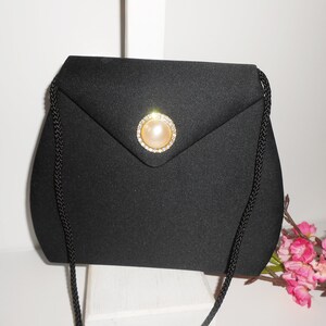May include: A black satin evening bag with a gold and rhinestone clasp featuring a faux pearl. The bag has a long black braided strap.
