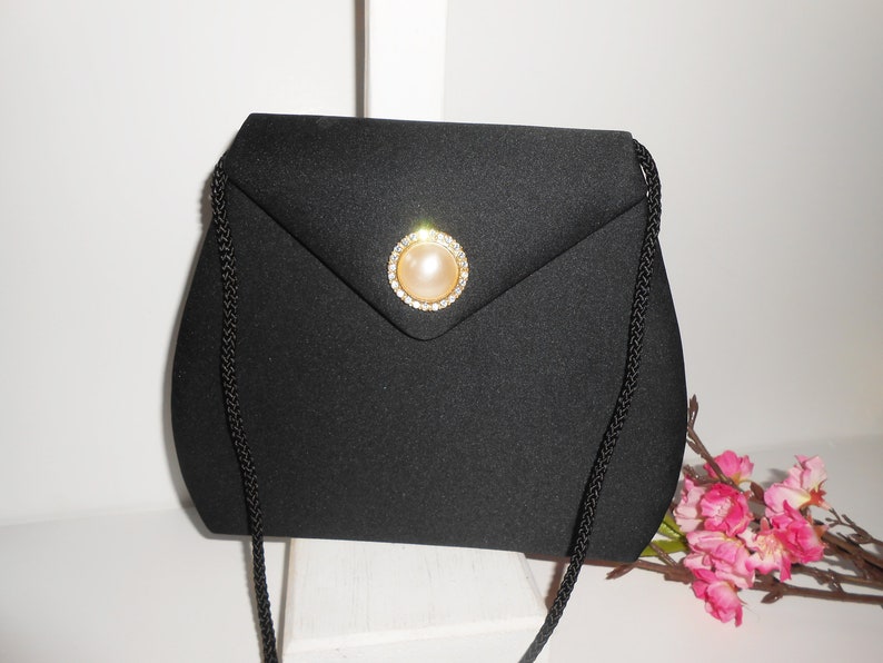 May include: A black satin evening bag with a gold and rhinestone clasp featuring a faux pearl. The bag has a long black braided strap.