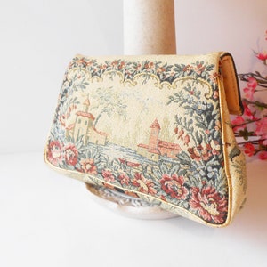 May include: A vintage tapestry clutch bag with a floral design and a scene of a castle by a lake. The bag is beige with pink, red, green, and blue accents.