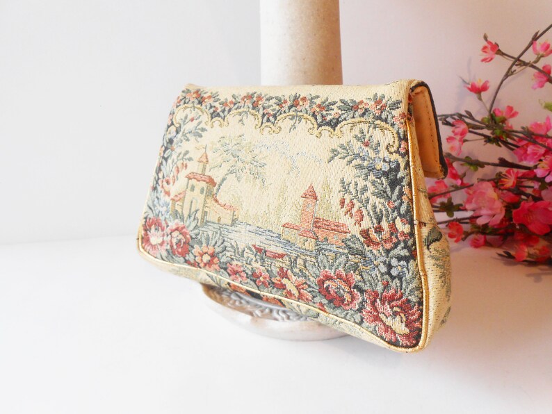 May include: A vintage tapestry clutch bag with a floral design and a scene of a castle by a lake. The bag is beige with pink, red, green, and blue accents.