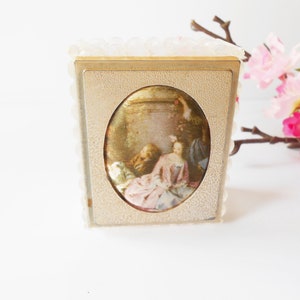 May include: A small, white, decorative box with a gold frame and a vintage oval picture of a woman in a pink dress. The box is surrounded by a white, beaded trim.