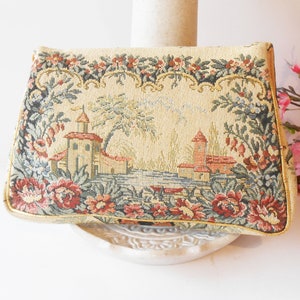 May include: A vintage tapestry clutch bag with a floral design and a scene of a small town with buildings and trees. The bag is beige with red, green, and blue accents.