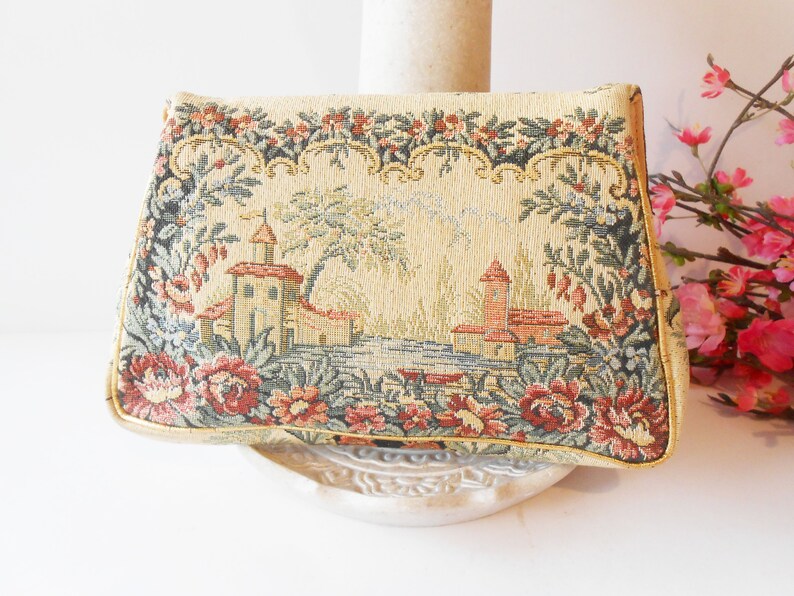 May include: A vintage tapestry clutch bag with a floral design and a scene of a small town with buildings and trees. The bag is beige with red, green, and blue accents.
