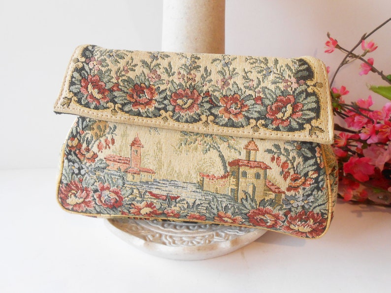May include: A vintage tapestry clutch bag with a floral and landscape design. The bag has a flap closure and a cream background with red, green, and brown accents.