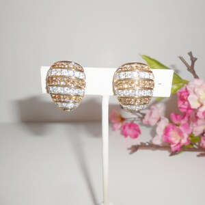 May include: A pair of gold-toned clip-on earrings. Each earring features a domed shape with alternating horizontal bands of clear and amber-colored crystals. The earrings are displayed on a white stand against a soft background.