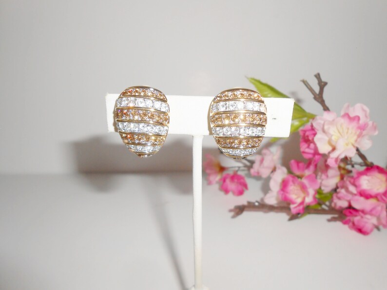 May include: A pair of gold-toned clip-on earrings. Each earring features a domed shape with alternating horizontal bands of clear and amber-colored crystals. The earrings are displayed on a white stand against a soft background.