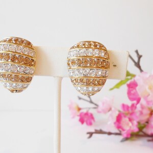 May include: A pair of gold-toned clip-on earrings. Each earring is oval-shaped and features alternating rows of clear and amber-colored rhinestones. The earrings are displayed on a white stand against a soft background.