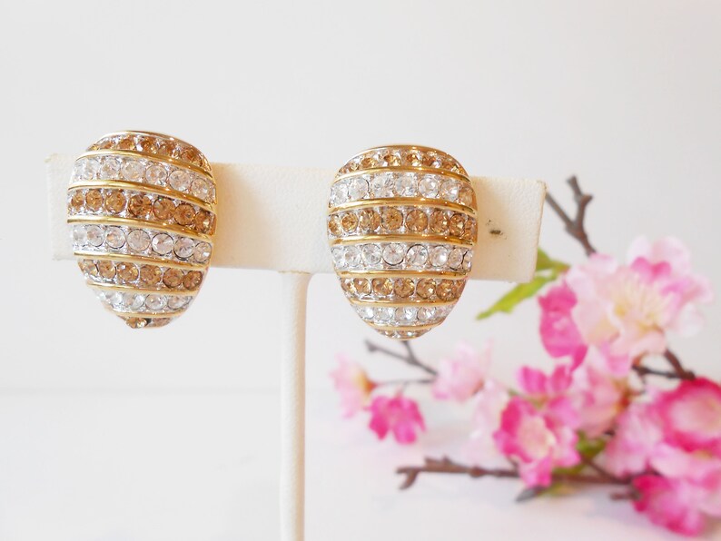 May include: A pair of gold-toned clip-on earrings. Each earring is oval-shaped and features alternating rows of clear and amber-colored rhinestones. The earrings are displayed on a white stand against a soft background.