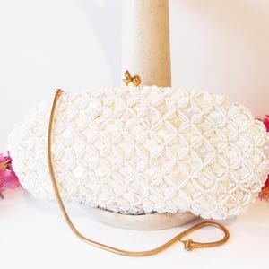 May include: Ivory beaded clutch bag with a gold chain strap. The bag features a geometric pattern of small beads and a gold clasp. The chain is a delicate, thin design. Pink flowers are visible in the background.