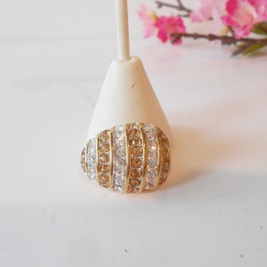 May include: An oval-shaped brooch with alternating rows of clear and amber-colored rhinestones set in a gold-tone metal. The brooch is displayed on a white stand, with pink flowers in the background.