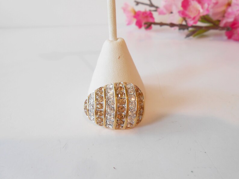 May include: An oval-shaped brooch with alternating rows of clear and amber-colored rhinestones set in a gold-tone metal. The brooch is displayed on a white stand, with pink flowers in the background.