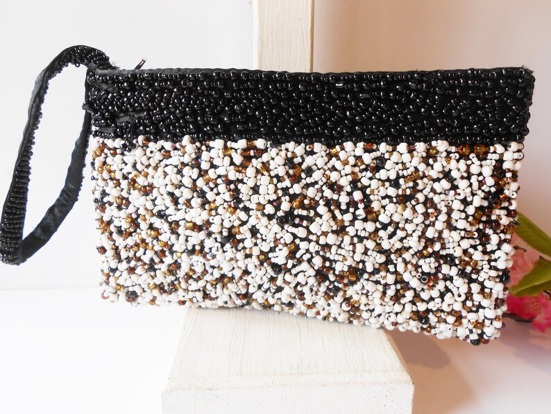 May include: A black and white beaded clutch with a black strap. The clutch is made of small, round beads in a variety of colors, including white, black, and brown.