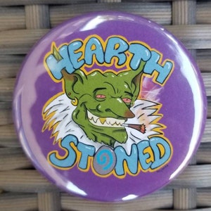 Puede incluir: Botón morado con un duende verde de dibujos animados fumando un porro. El duende tiene los ojos rojos y una sonrisa de oreja a oreja. El texto del botón dice "HEARTH STONED" en azul y amarillo.