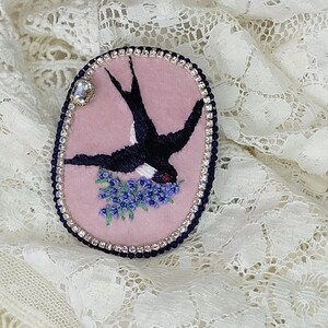 May include: A pink felt brooch with a black embroidered swallow carrying a sprig of purple flowers. The brooch is surrounded by a row of clear rhinestones.