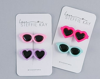 May include: Four hair clips shaped like heart-rimmed sunglasses in pink, lavender, hot pink, and light blue, each with black lenses. The clips are mounted on two white display cards labeled Love STEFFIE KAY with the handle @lovesteffiekay.