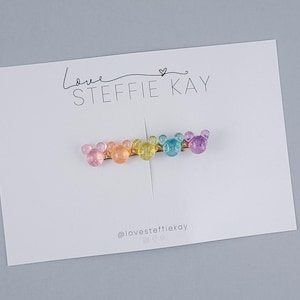Puede incluir: Una pinza para el pelo de color arcoíris con cinco cuentas en forma de Mickey Mouse. La pinza se muestra en una tarjeta blanca con el texto "Love STEFFIE KAY" y el identificador de Instagram "@lovesteffiekay".