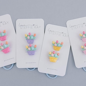 Könnte beinhalten: Vier Karten mit dekorativen Blumen-Anstecknadeln. Jede Karte zeigt zwei Anstecknadeln in Form von Blumentöpfen mit bunten Tulpen. Die Anstecknadeln sind rosa, lila und gelb. Die Karten haben eine blaue Schlaufe.