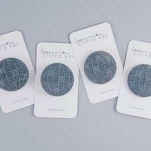 May include: Four gray globe-shaped accessories on white cards. Each globe has a grid pattern. The cards have the text "Love Steffie Kay" and a small heart. The background is a light gray.