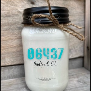 May include: A white mason jar candle with a black lid and twine bow. The candle is labeled with the text "06437 Guilford. Ct." and "16 oz./454 (g) hand-poured soy 80+ hour burn-time beanwick.com".