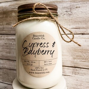 Puede incluir: Una vela blanca de soja en un tarro de vidrio transparente con tapa marrón y un lazo de cuerda marrón. La etiqueta dice "Beanwick Candle Co. Cypress & Bayberry" e incluye detalles como "Peso neto 16 oz. 454 (g)", "Tiempo de combustión de más de 80 horas", "100% soja", "Hecho a mano en CT." y "www.beanwick.com".