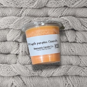 Puede incluir: Un pequeño recipiente de plástico transparente que contiene una cera de soja perfumada de color naranja. La etiqueta dice "Maple pumpkin Crunch" y "Beanwick Candle Co.". Sobre una tela gris de punto de cable.