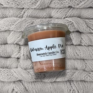 Op de afbeelding: Een kleine, bruine sojawas smelt in een doorzichtige plastic container met een wit label waarop "Warm Apple Pie" en "Beanwick Candle Co. 1 oz. Soy Wax Pop (melt)" staat.