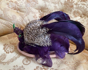 May include: A purple and gray feathered hat with a pink base. The hat is decorated with green feathers.