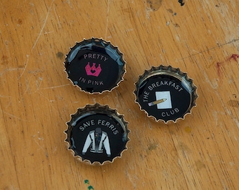 May include: Three black bottle caps with different designs. The first cap says "Pretty in Pink" with a pink crown. The second cap says "The Breakfast Club" with a pencil and paper. The third cap says "Save Ferris" with a person wearing a jacket.