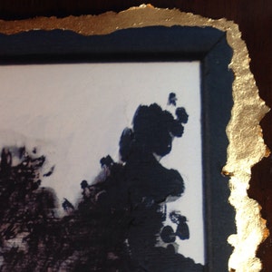 May include: A close-up of a framed artwork with a gold leaf border. The artwork features a black and white abstract design with a white background.