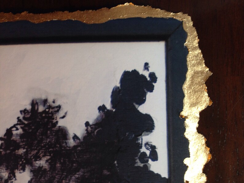 May include: A close-up of a framed artwork with a gold leaf border. The artwork features a black and white abstract design with a white background.