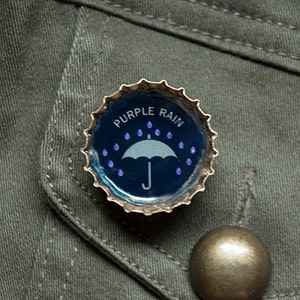 Könnte beinhalten: Ein lila Flaschenverschluss mit einem weißen Regenschirm und dem Aufdruck "Purple Rain".