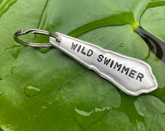Op de afbeelding: Een sleutelhanger in de vorm van een zilveren lepel met de woorden "WILD SWIMMER" erop gegraveerd. De sleutelhanger ligt op een groen blad met waterdruppels.