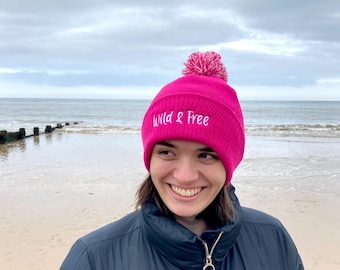 Op de afbeelding: Een roze beanie met een witte pompon en de tekst "Wild & Free" geborduurd op de voorkant. De beanie wordt gedragen door een persoon op een strand.