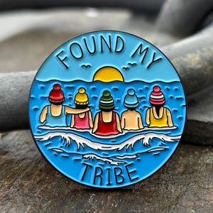 Puede incluir: Un pin redondo de esmalte con un fondo azul que representa cinco figuras en el agua con gorros de punto de colores. El pin tiene las palabras "FOUND MY TRIBE" en letras blancas, con un sol amarillo y pájaros en el cielo.