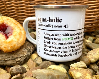 Op de afbeelding: Witte emaille mok met een zilveren rand, met het woord "aqua-holic" en een definitie. Een koekje met rode vulling ligt naast de mok op stenen. De definitie van de mok bevat de term "FOMO".