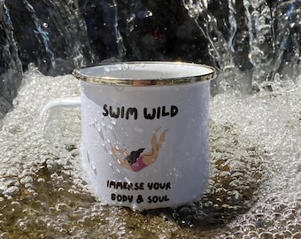 Op de afbeelding: Een witte emaille mok met een gouden rand. De mok heeft een zwart-witte illustratie van een zwemmend persoon met de tekst "Swim Wild" en "Immerse your body & soul".