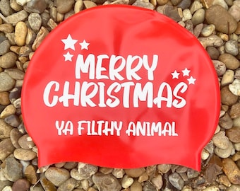 May include: A bright red swim cap with the text "MERRY CHRISTMAS YA FILTHY ANIMAL" in white, along with white star graphics. The cap is displayed on a bed of small, smooth stones.