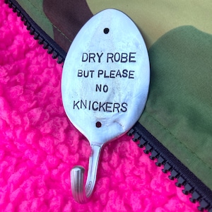 May include: A silver spoon shaped hook with the text "DRY ROBE BUT PLEASE NO KNICKERS" engraved on it. The hook is hanging on a pink fuzzy surface.