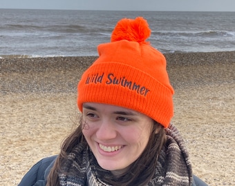 May include: Orange knit beanie with a black pom pom and the text "Wild Swimmer" embroidered on the front.