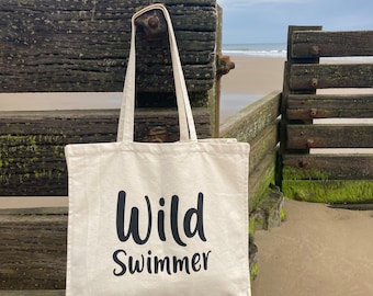 May include: A white canvas tote bag with black text that reads "Wild Swimmer". The bag is sitting on a wooden bench with a beach in the background.