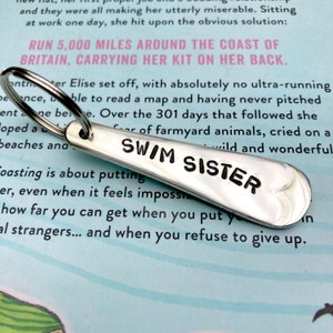 Op de afbeelding: Zilveren sleutelhanger met een peddelvormig label met de tekst "Swim Sister" in zwarte letters.