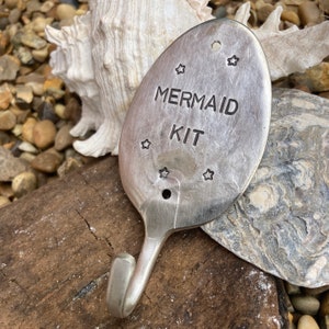 May include: A silver spoon with a hook on the end. The spoon has the words "Mermaid Kit" engraved on it, along with several stars.