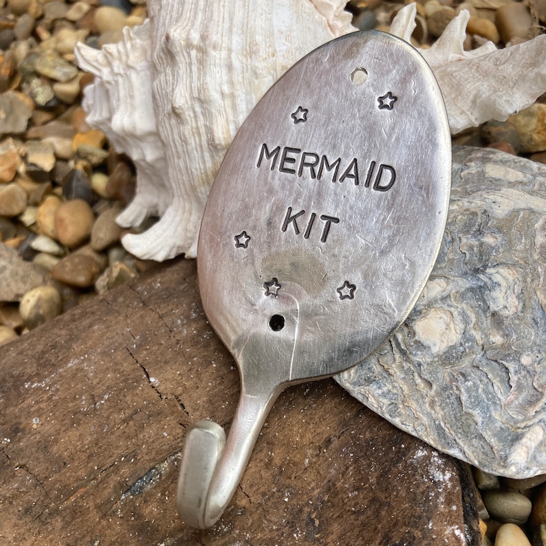 May include: A silver spoon with a hook on the end. The spoon has the words "Mermaid Kit" engraved on it, along with several stars.