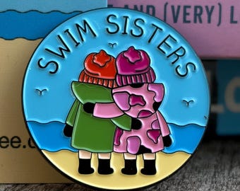 Op de afbeelding: Een ronde emaille pin met de tekst "SWIM SISTERS". De pin toont twee figuren die elkaar omarmen, gekleed in jassen en hoeden. De achtergrond toont een strandscène met blauw water en geel zand.