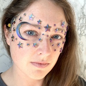 May include: A woman wearing a sheer face mask with iridescent stars and a crescent moon design. The mask is made of a fine mesh material and is decorated with holographic stars and a crescent moon.