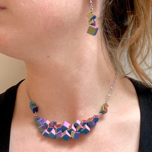 May include: A geometric jewelry set featuring a necklace and earrings. The necklace and earrings are composed of small, colorful cube-shaped beads in shades of blue, purple, pink, and gold. The necklace has a silver chain.
