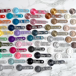 May include: A collection of 36 small, round, colorful buttons with various names written on them. The buttons are arranged in rows and columns on a white surface.