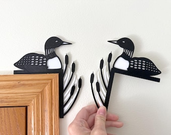 May include: Black metal loon and cattail corner decor. The loon has a white chest and detailed wing design. The cattails are white and black. The decor is designed to be placed on a corner.