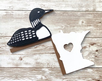May include: A black and white loon cutout with a brown base, paired with a white cutout of the state of Minnesota, featuring a heart-shaped cutout. The items are placed on a light wood grain surface.
