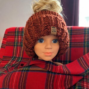 May include: A rust-coloured chunky knit beanie with a high opening at the crown for a ponytail. The hat features a textured ribbed pattern and a small rectangular tan leather tag on the front with the text MADE WITH LOVE in black ink.